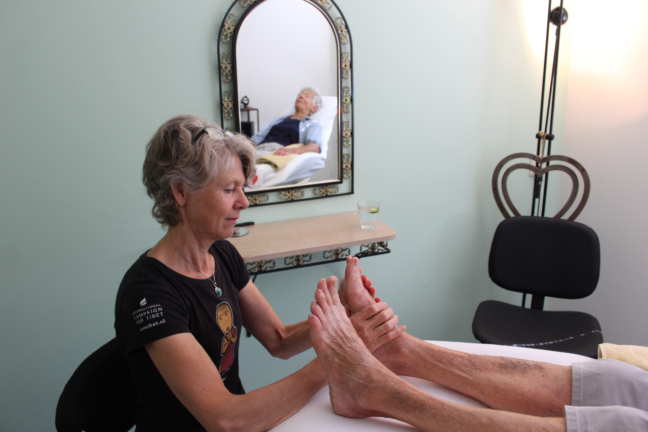 Voetreflexologe Carolinda van der Linden masseert een paar voeten. In de spiegel is het gezicht van de cliënt zichtbaar.